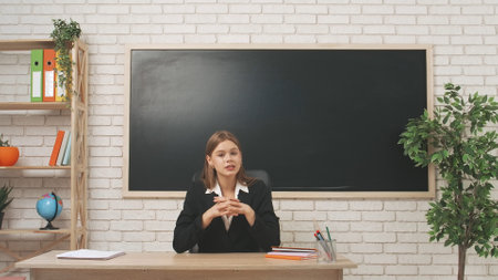 Young woman college teacher sitting at desk in classroom in front of chalkboard explains lesson to...