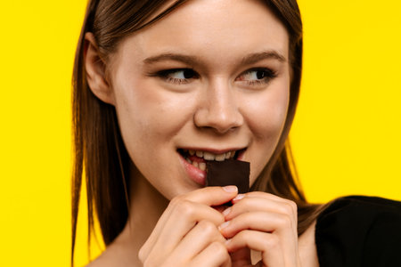 Woman enjoying a delicious piece of chocolate on a vibrant yellow backgroundの写真素材
