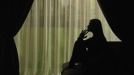 Silhouette of a distressed woman in front of backlit curtainsの写真素材