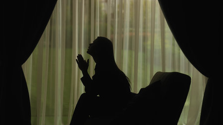 Silhouette of a woman praying in a pensive pose in the light of the windowの写真素材