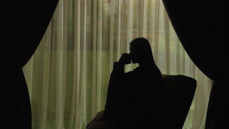Silhouette of a distressed woman in front of backlit curtainsの写真素材