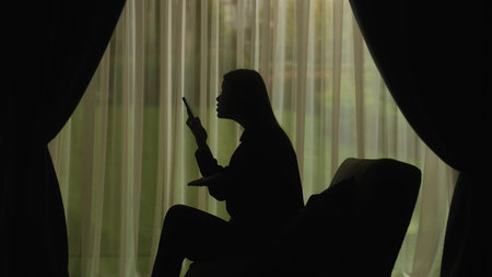 Silhouette of a Person Using Smartphone by Window Curtainsの写真素材