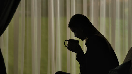 Silhouette of a female, distressed sitting on a sofa and holding a cup of hot tea.の写真素材