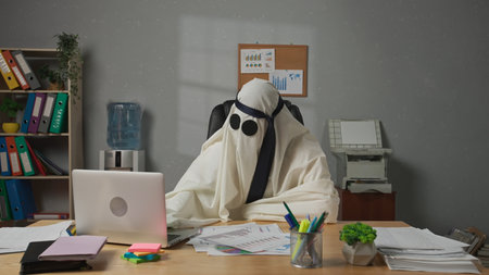 Halloween businessman ghost in white sheet and necktie over head sits at office desk works on laptop, satisfied expression. Happy halloween business concept.の写真素材