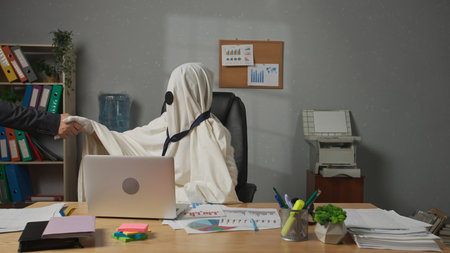 Halloween businessman ghost in white sheet sits at office desk works on laptop, meets with partner...