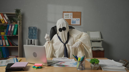 Halloween businessman ghost in white sheet sits at office desk dancing in chair and catching flying paper documents in reverse. Happy halloween business concept.の写真素材