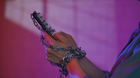 Shot showing a woman hands restrained by a metal chain with a mobile phone, symbolizing...
