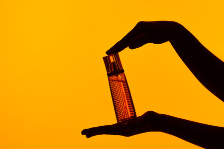 Silhouetted Hand Holding Elegant Glass Perfume Bottle on Orange Backgroundの写真素材