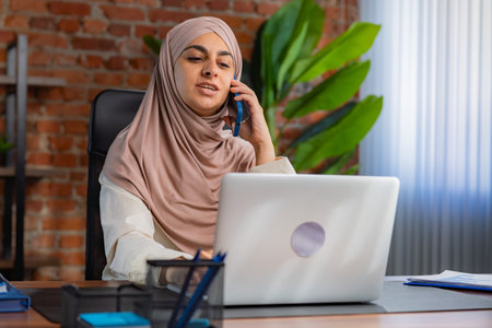 Focused Professional Woman in Hijab Using Phone and Laptop at Office Deskの写真素材