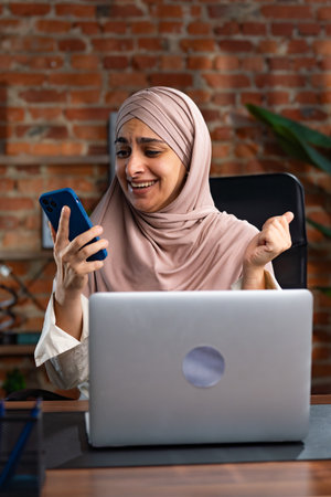 Smiling Woman in Hijab Using Smartphone at Modern Workspaceの写真素材