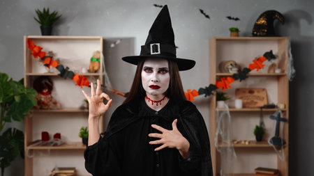 A vampire witch in a black cloak and hat makes an OK gesture with her fingers
