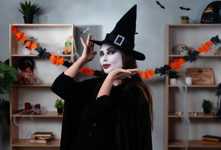 A vampire witch in a black cloak and hat poses standing in a room decorated for Halloween