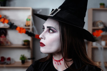 A vampire witch in a black cloak and hat poses standing in a room decorated for Halloweenの写真素材