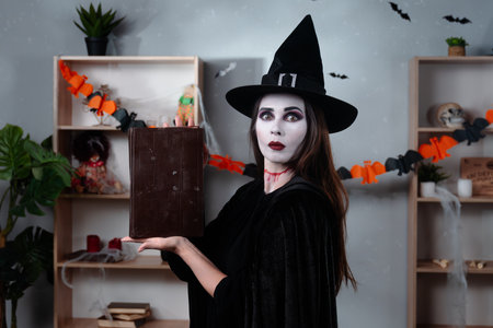 A vampire witch in a black cloak and hat shows off a book of magic while standing in a room decorated for Halloween.の写真素材