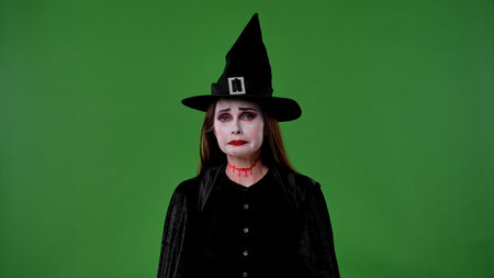 A witch with vampire makeup, dressed in a black cloak and hat, looks sadly into the camera