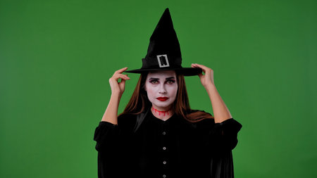 A witch with vampire makeup, in a black cloak and hat, looks at the camera and adjusts her hat