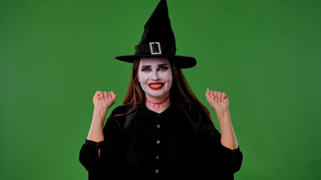 A witch with vampire makeup in a black cloak and hat, poses cheerfully