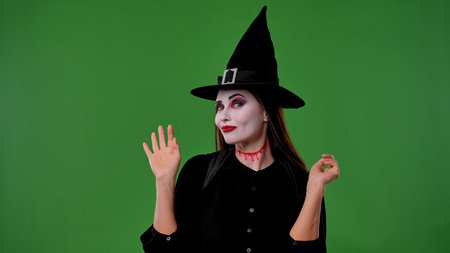 A witch with vampire makeup, dressed in a black cloak and hat, waves hello