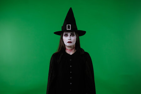 A witch with vampire makeup, wearing a black cloak and hat, looks at the camera and appears very...