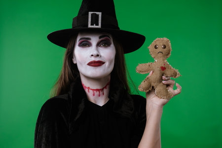 A witch with vampire makeup, wearing a black cloak and hat, poses with a voodoo doll and a needle...