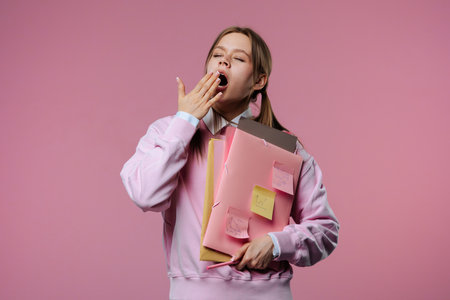 Tired young female student yawns while holding folders and notesの写真素材