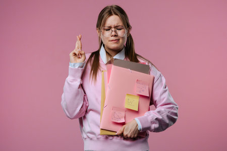 A young female student crosses her fingers for good luck, holding folders and notes in her handsの写真素材
