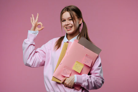 A young female student makes an OK gesture while holding folders and notesの写真素材