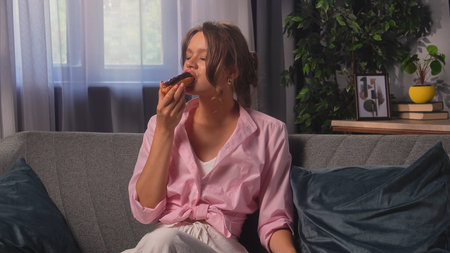 Young Woman Enjoying a Chocolate Glazed Eclair at Home on a Cozy Sofa