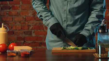 A chef wearing black gloves slices fresh cucumbers on a wooden board in a rustic kitchen setting. The process of preparing food in a cafe or restaurant.の写真素材