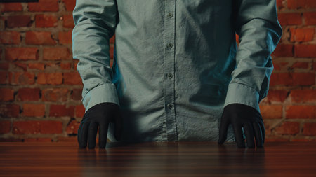 A person in a blue shirt and black gloves stands with hands on a wooden table against a brick wall.の写真素材