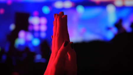 Clapping hands in red light against a blue bokeh background at a concert. Festive atmosphere for a party, event, New Years Eve, and Christmas.の写真素材