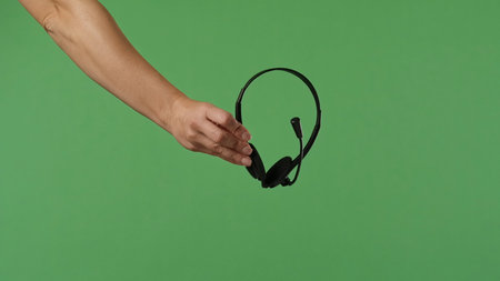 A woman hand holding a black headset on a green chroma key background. Shot for a design related to office work and the provision of services and call center support.の写真素材
