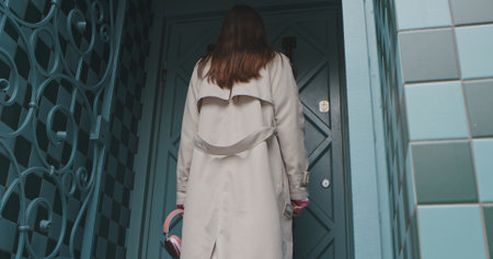 A woman in a beige trench coat stands facing a blue door, holding pink headphones in her hand.の写真素材