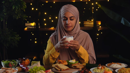 A Muslim woman wearing a hijab sits at a festive table laden with various dishes, holding a glass of...