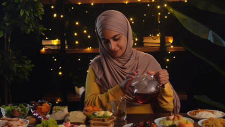 A muslim woman in a hijab pours tea during a cozy dinner setting with food, surrounded by warm fairy lights. Traditional Eastern dishes are laid out on a table in a restaurant or cafe.の写真素材