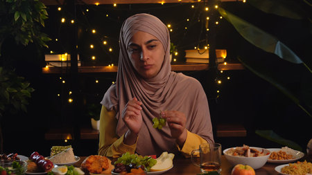 A muslim woman in a hijab is seated at a table with a variety of dishes, holding grapes, with warm lights in the background.の写真素材