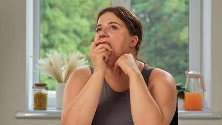 A woman in a gray tank top eats an apple while seated at a table in a bright and airy room.