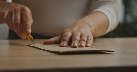 Hands woman cutting a piece of cardboard with a utility knife on a wooden table. Careful craftsmanship in focus.の写真素材