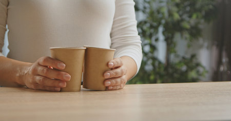 Close up of woman hands holding two recyclable paper coffee cups on a wooden table with green plant...