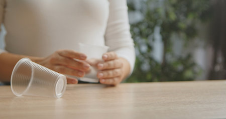 A woman holds a transparent plastic cup, with another cup lying on a wooden table in a blurred background.の写真素材