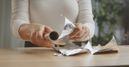 Close up of woman hands manipulating a shiny roll of aluminum foil on a wooden table, highlighting household activity.の写真素材