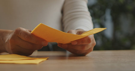 Close up view of woman hands carefully folding a yellow paper napkin on a wooden surface, showing detail.の写真素材
