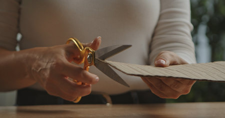 A close up image showing woman hands using gold scissors to cut a piece of brown corrugated cardboard on a table.の写真素材