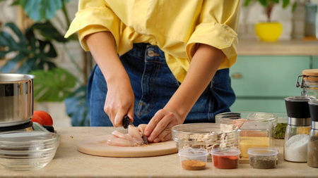 A close up shot of a woman slicing raw chicken breast on a cutting board, surrounded by spices and vegetables. Preparing lunch or dinner in a home kitchen.の写真素材