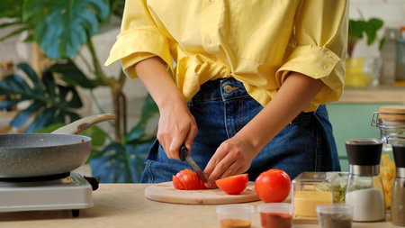 Close up of woman in yellow shirt slicing tomato on a kitchen counter with utensils and greenery. Preparing lunch or dinner in a home kitchen.の写真素材