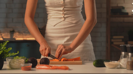 A woman prepares healthy food in her kitchen and cuts carrots into strips. Cucumbers, avocados, fresh lettuce leaves, salmon, and radishes are laid out on the table.の写真素材