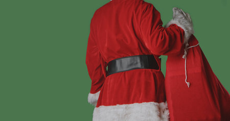Rear view of Santa in a red suit holding a large bag of gifts against a green background, symbolizing Christmas deliveries, holiday traditions, and the festive season.の写真素材