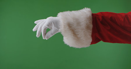 Santa hand in a red suit with a white fur cuff and glove isolated on a green background. Perfect for...