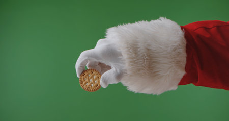 Santa Claus hand in a red suit with a white fur cuff and glove holds a round holiday cookie, isolated on a green background. Perfect for Christmas banners, cards, and holiday advertising.の写真素材