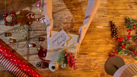Top view of women hands holding a Christmas gift on a wooden table, wrapped in kraft paper with...